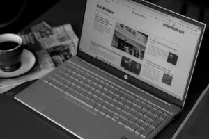 Home Black and white image of a laptop displaying news articles, accompanied by a cup of coffee and newspapers.